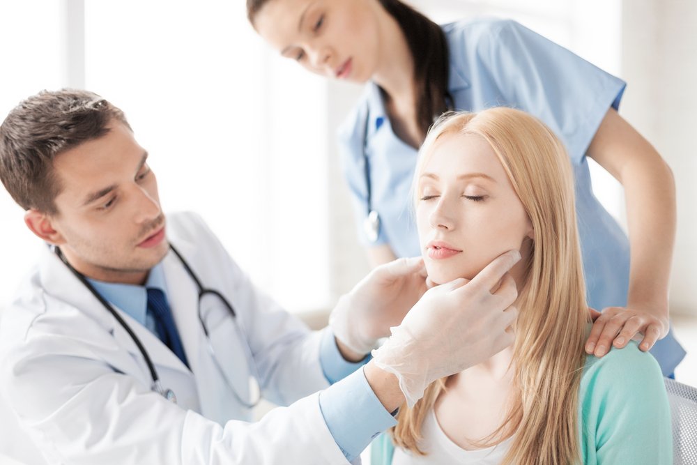 Doctor wearing gloves examining the face of a female patient with a nurse standing behind her in an examination room